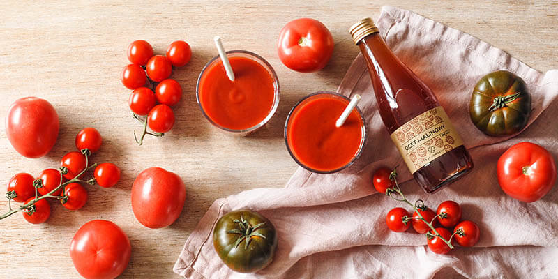 na drewnianym stole leży butelka octu malinowego Racjonalni, obok stoją dwie szklanki z sokiem pomidorowym i octem malinowym, na blacie leżą rozrzucone pomidory