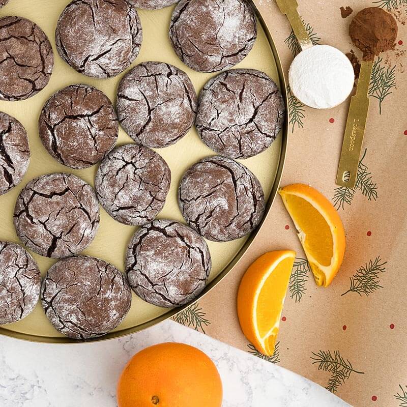 świąteczne, popękane ciasteczka czekoladowe na złotej tacy; w tle świąteczny papier do pakowania prezentów, miedziane miarki z cukrem pudrem i kakao, pomarańcza