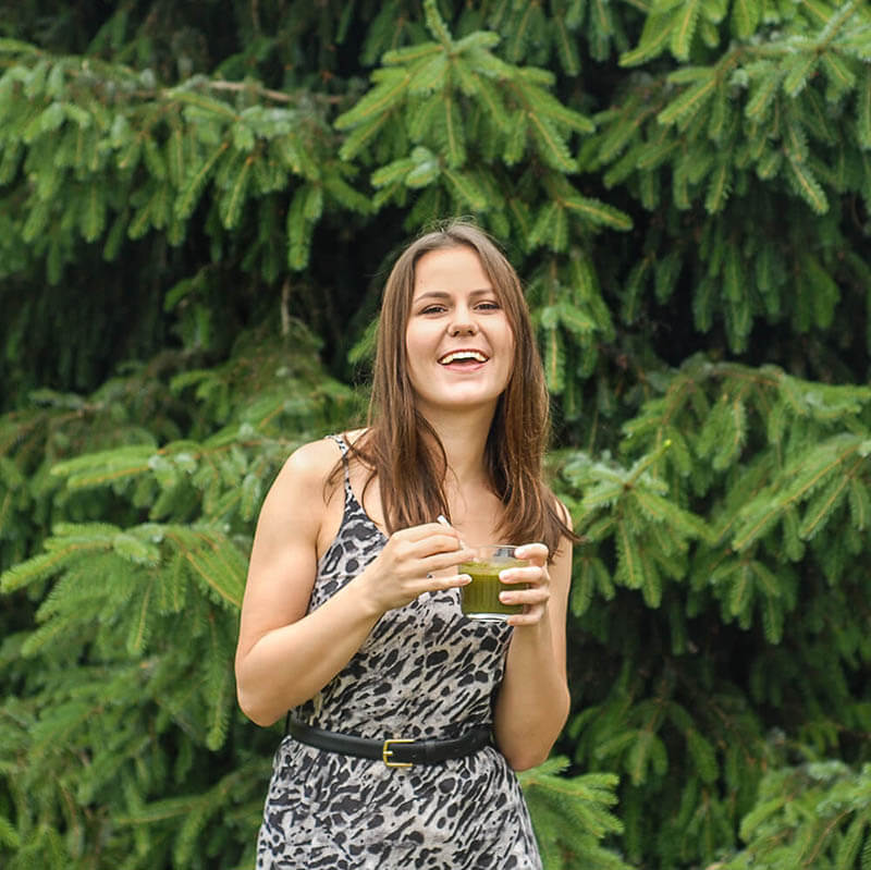 uśmiechnięta dziewczyna w letniej sukience trzyma szklankę i pije sok z trawy pszenicznej; na tle drzew iglastych